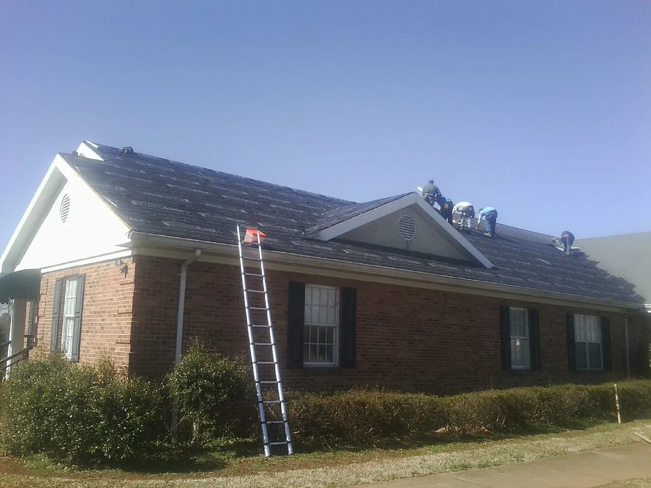 Crew tearing shingles off a brick ranch roof with a ladder set — Emergency Roof Repair work by our crew in New Galilee