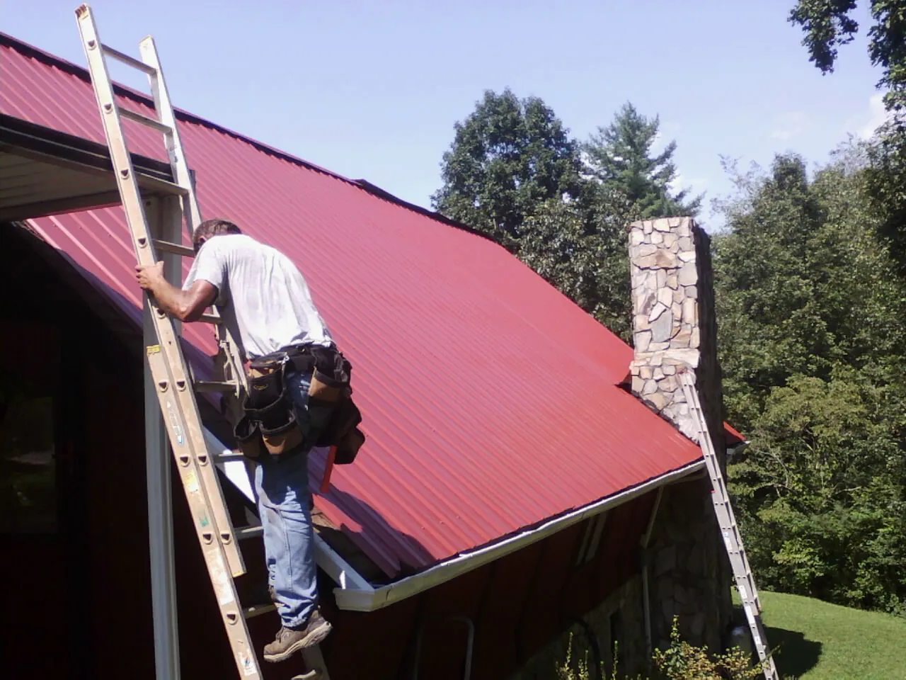 Roofer on a ladder beside a red metal roof and stone chimney — Soffit & Fascia Repair by Roofing Brothers in New Galilee